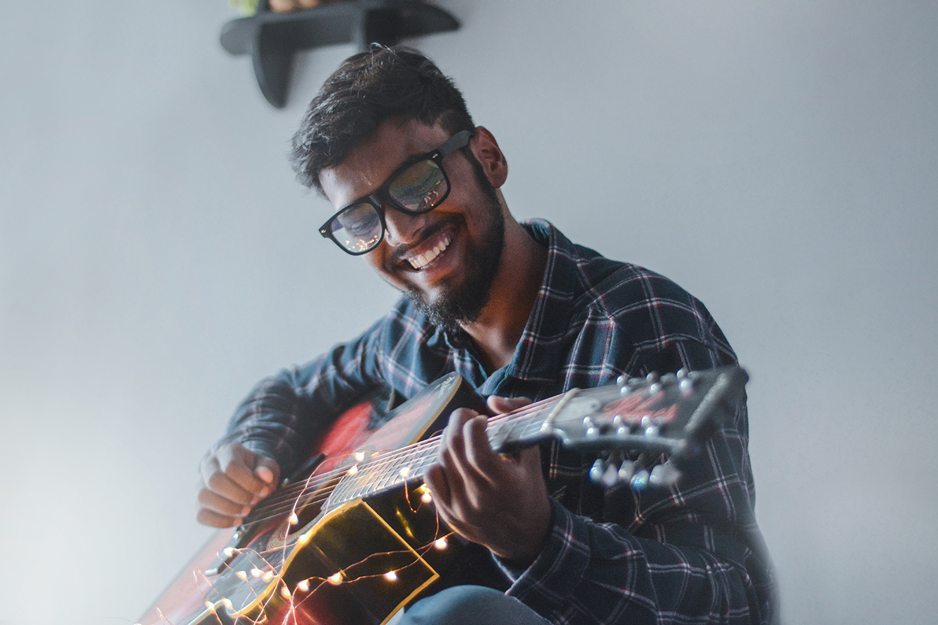 A young South Asian man plays a guitar with a broad smile on his face. 