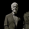 Black and white photo of Professor Dick Hobbs wearing a suit standing against a brick and concrete wall. 