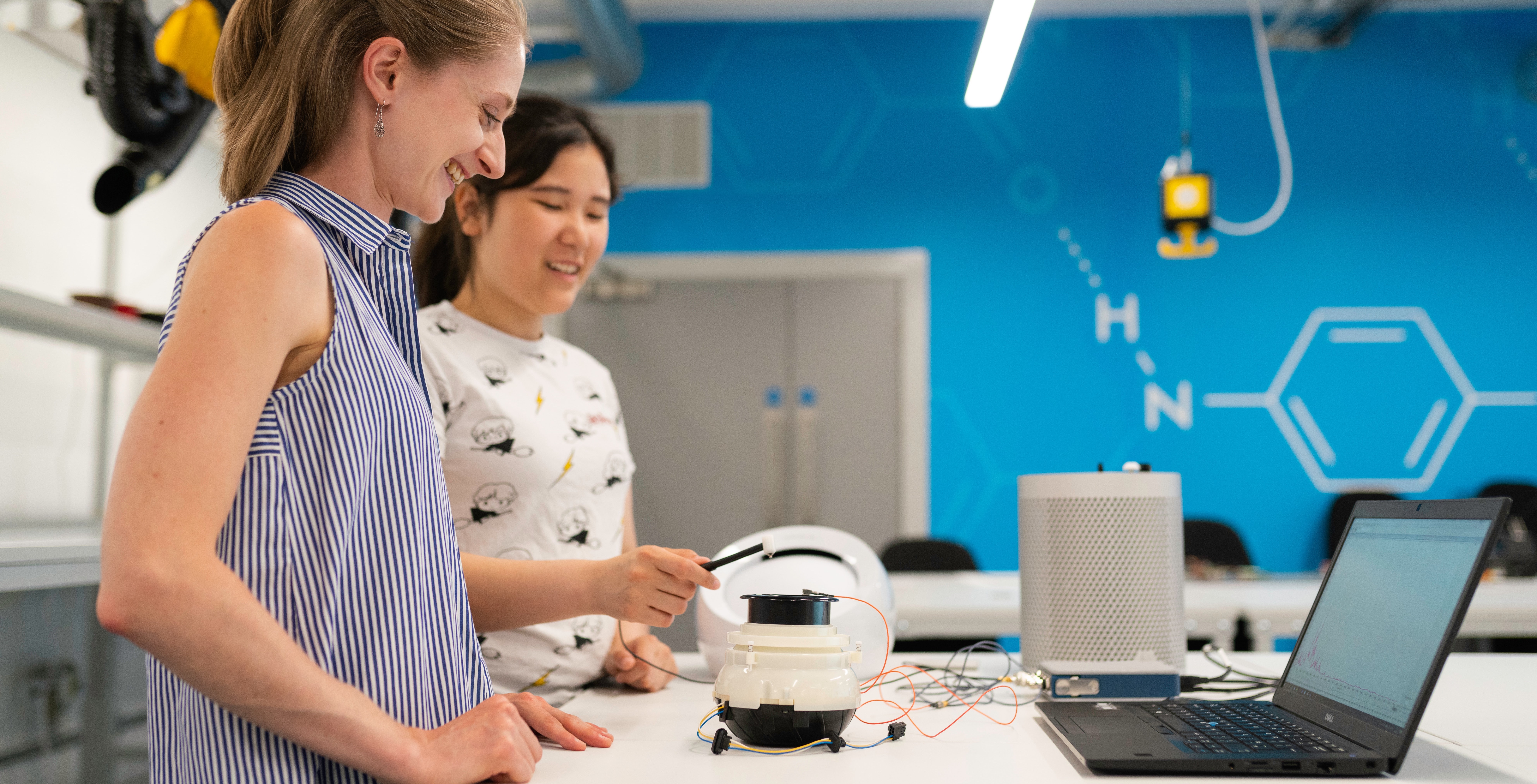 2 women in an engineering lab, standing over a technical device having a conversation 