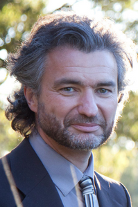 Adam wears a blue and grey suit. He is standing against a natural leafy background. He looks at the camera. 