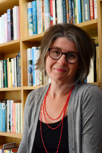 Tegan wears a black singlet, a grey long-sleeved cardigan, and several bright red necklaces. She has short brown and blonde hair, and wears rounded brown glasses. 