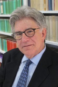 Dennis wears a suit and tie. The suit is black while the tie and shirt are blue. He has grey hair, and wears black framed glasses. He is sitting in front of a bookshelf. He looks into the camera and smiles slightly.