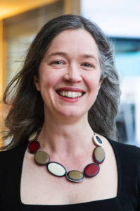 Kate wears a square necked black top, with a colourful necklace. Her long hair is swept back over her shoulders. She smiles at the camera. 