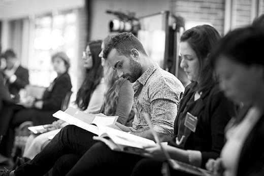 A black and white photo of the audience listening to the presentations with a focus on two people who are looking at their booklets.