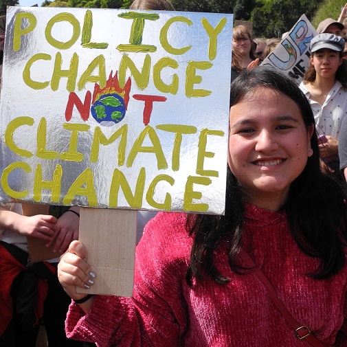A young person in amongst a crowd holds a sign saying 'policy change not climate change'. 