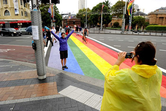 A road crossing is painted in purple, blue, green, yellow, orange and red stripes. A woman stands on the crossing posing for the camera while another takes her photo.