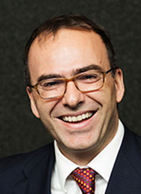 Dimitris wears a suit with a red tie. He has glasses. 