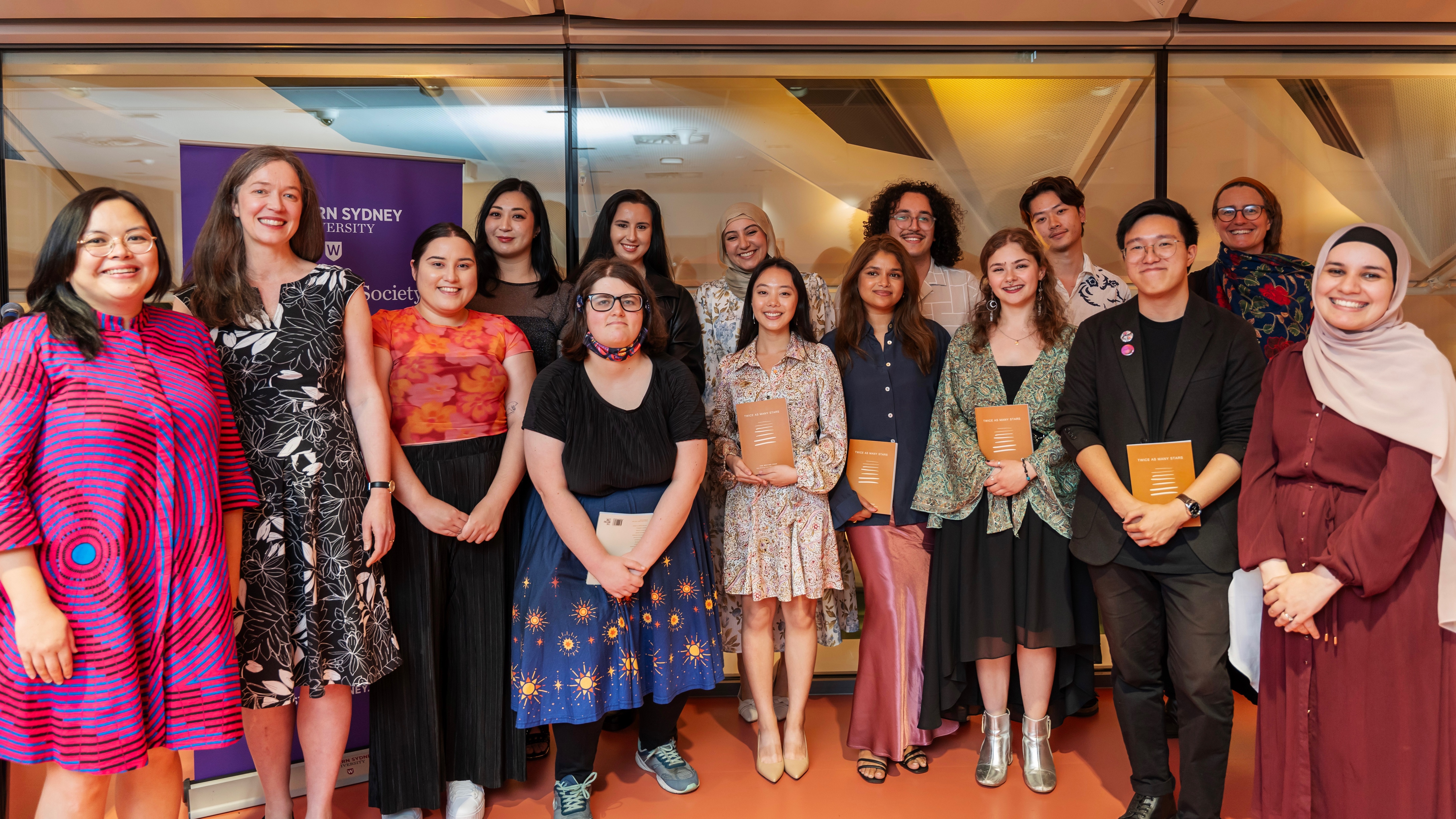 A group of writers, mentors, and WSRC staff stand together and smile. The shot is full length, and many of the people in the front row hold a copy of Twice as Many Stars.