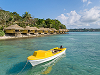 A small yellow and white boat floats in the clear blue water by the shore in Vanuatu. There are huts on the shoreline and trees on the hillside behind them.