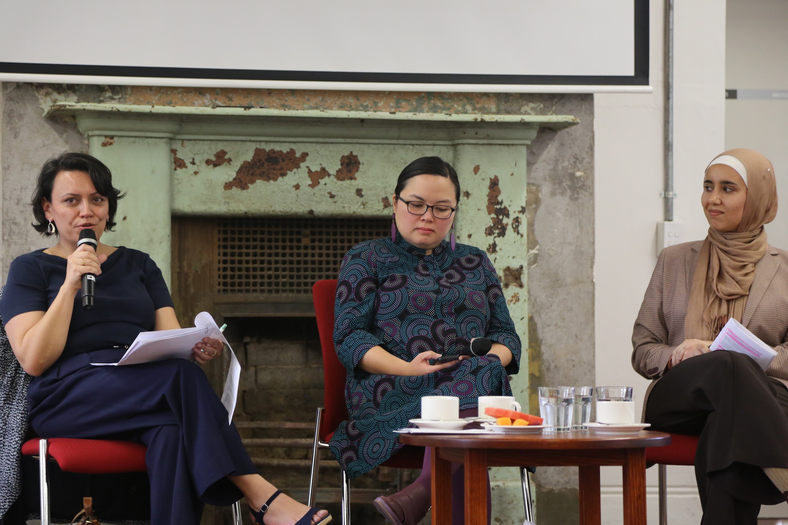 Three women sit in front of a green fireplace. The woman on the left is speaking into a microphone and is addressing the audience off camera. The woman in the middle wears a dark blue patterned dress, and has thin-framed black glasses. The women on the left wears a beige and cream coloured hijab and top, and holds a stack of papers.