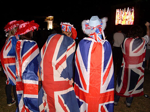The backs of people who are wrapped in the UK flag, as they stand watching the closing ceremony on a TV screen in a park.