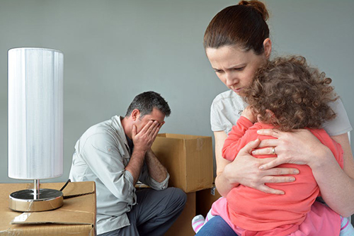 A woman hugs a child with a man in the background holding his head in his hands. Boxes are in the background.
