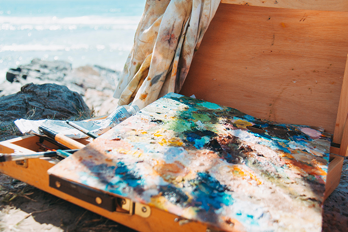 An abstract painting on canvas and a wooden box of art supplies sits on the rocks near the ocean.