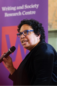 Sandra wears a long sleeved black top. She holds a microphone and is addressing an audience during a talk.