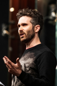 Ben wears a black sweater. He has curly brown hair and a moustache and beard. He is looking off-camera, speaking to an audience. 