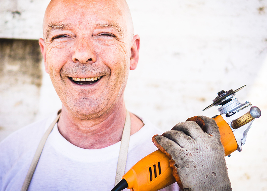 An older white man, holding an angle grinder, smiles broadly