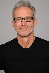 Chris Conti wears a black shirt. He smiles at the camera. He has grey short hair and black rectangle glasses.