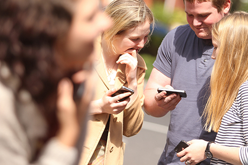 A group of young people standing together holding and looking at their mobile phones.