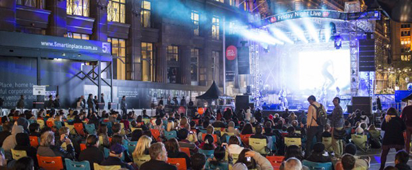 A photo of an outdoor concert next to city buildings with people seated facing the stage.