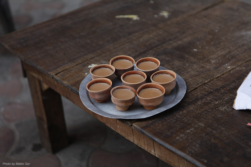 8 small clay teacups with tea in them sit on a round metal plate on a wooden table.