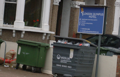A sign outside a hotel reads 'London Olympus hotel'. There are overflowing rubbish bins on the street directly outside.