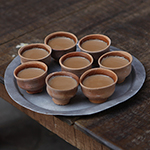 8 small clay teacups with tea in them sit on a round metal plate on a wooden table. 