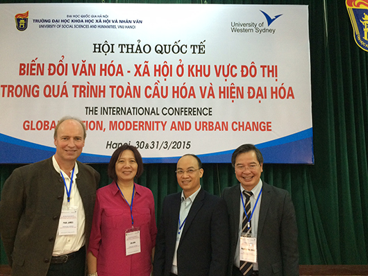 Paul James, Ien Ang, Pham Quang Minh and Nguyen Van Suu stand together at the conference in front of a green curtain and a conference sign.