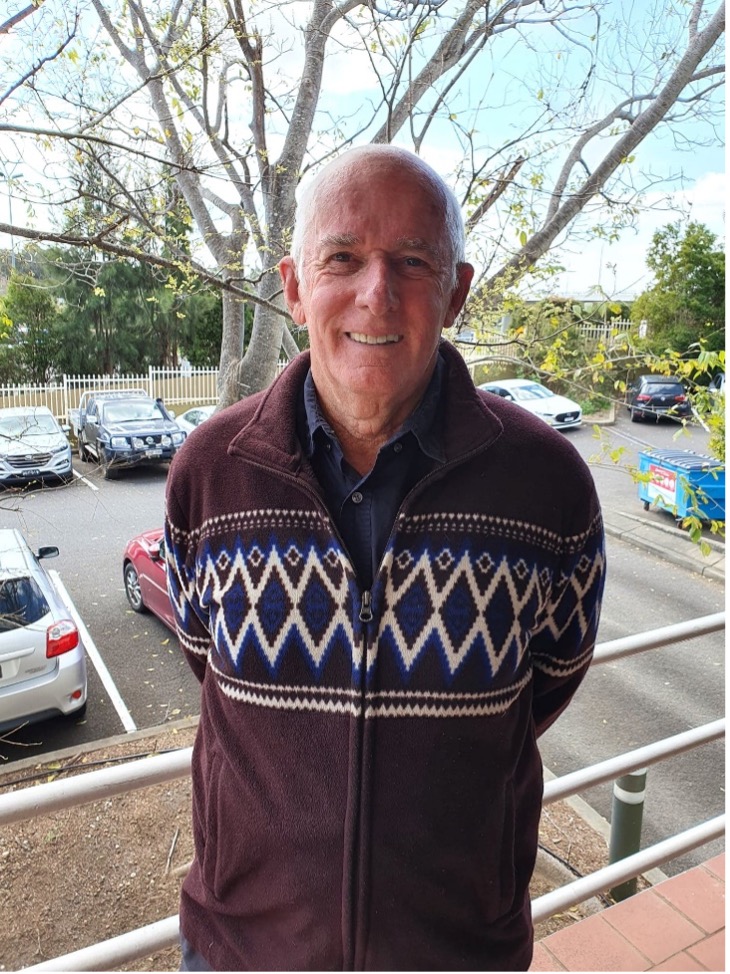 An image of an older man in a warm jacket smiling at the camera, with a bare tree and carpark behind him