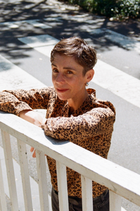 Catriona stands with her hands folder, leaning on a white fence. She wears a brown shirt and has short cropped hair.