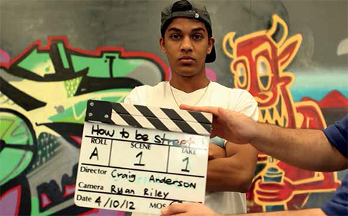 A young man stands behind a clapperboard and in front of a mural