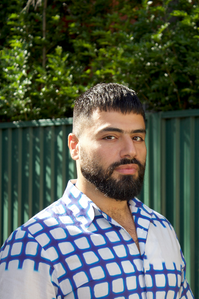 George Haddad stares into the camera. He is wearing a checkred white and blue shirt and stands in front of a green gate and tree.