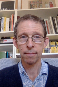 Chris Andrews sits on a grey chair in front of a bookshelf. He's has glasses, and wears a blue pattern shirt and dark blue cardigan.