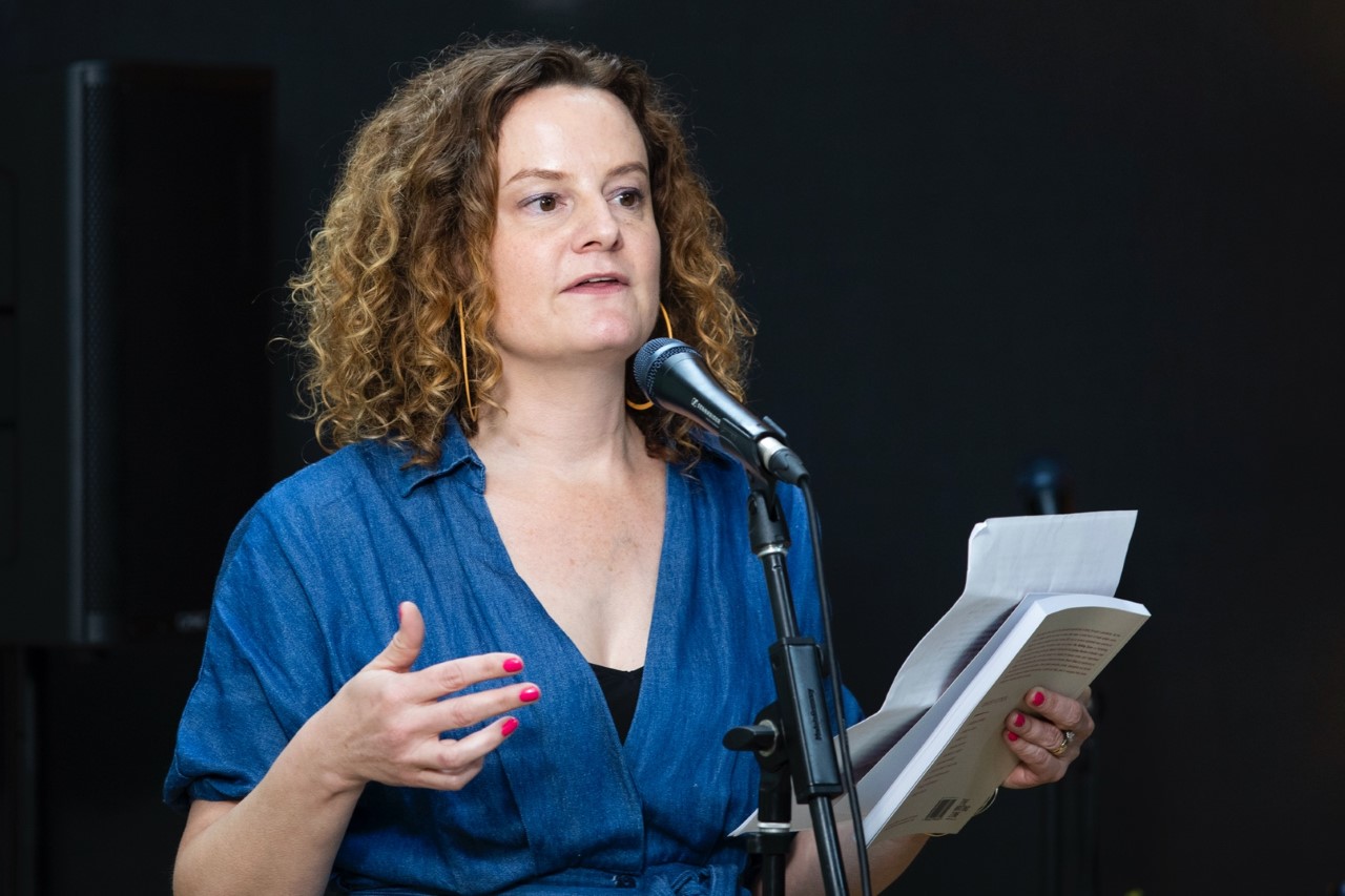 Felicity Castagna wears a blue shirt and holds a sheet of paper and book in her left hand. She is standing in front of a microphone, and speaking to an audience off screen. 