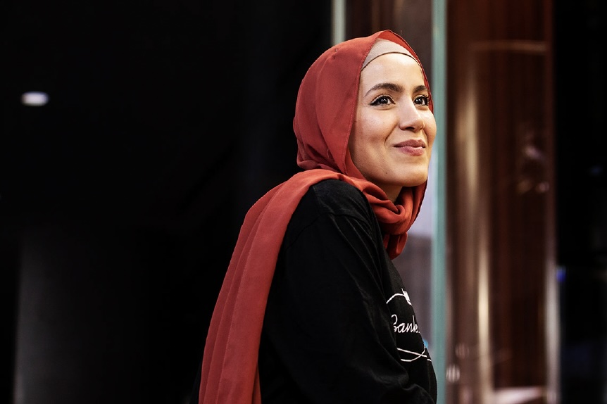 Sara Mansour is smiling at someone off camera. She wears a long-sleeved black jumper, and a orange-red hijab. 