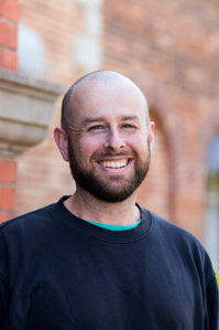 James is smiling at the camera. He is bald, and has brown stubble and a moustache. He wears a dark blue sweater with a green shirt underneath.