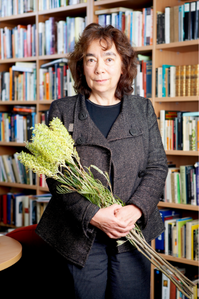 Alexis Wright holds a bouquet of native flowers. She stands in front of a bookshelf. She wears a grey jacket and navy blue shirt. She is smiling slightly at the camera.