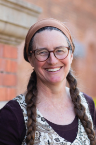 Melinda has long brown hair, tied into two plaits that lay down over her shoulders. She wears brown round glasses and an orange headband. She is smiling at the camera. 