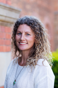 Lorraine has curly medium length blonde and brown hair. She wears a lightweight white blouse and a necklace with a green gemstone on it. She smiles at the camera., 