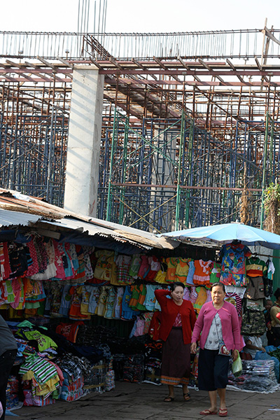 Two women walk past a market stall of children's clothing. It has a tin roof and umbrella over it. Behind the stall is scaffolding.