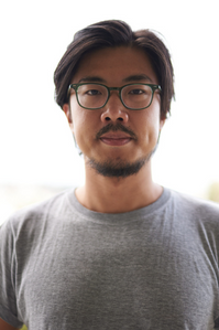 James Jiang is wearing a grey shirt and black glasses. He has beard and stubble, and is of East Asian descent. He is staring directly into the camera.