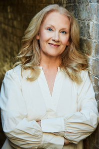 Mette wears a white blouse, and leans against a brick wall. her medium length blonde hair lays on her shoulders. She crosses her arms, and smiles at the camera.