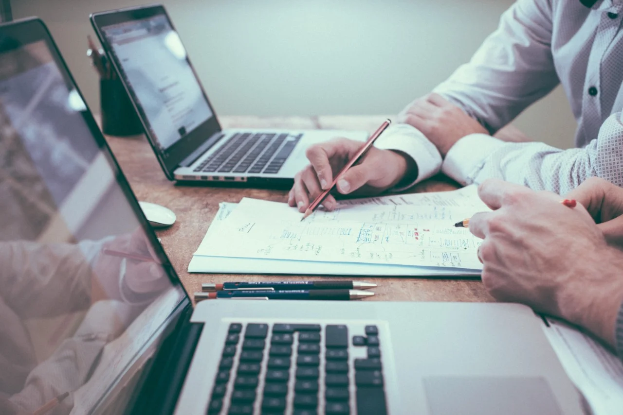 Hands shown doing paperwork near laptops