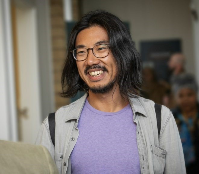 James Jiang is wearing a grey shirt and black glasses. He has beard and stubble, and is of East Asian descent. He is staring directly into the camera.