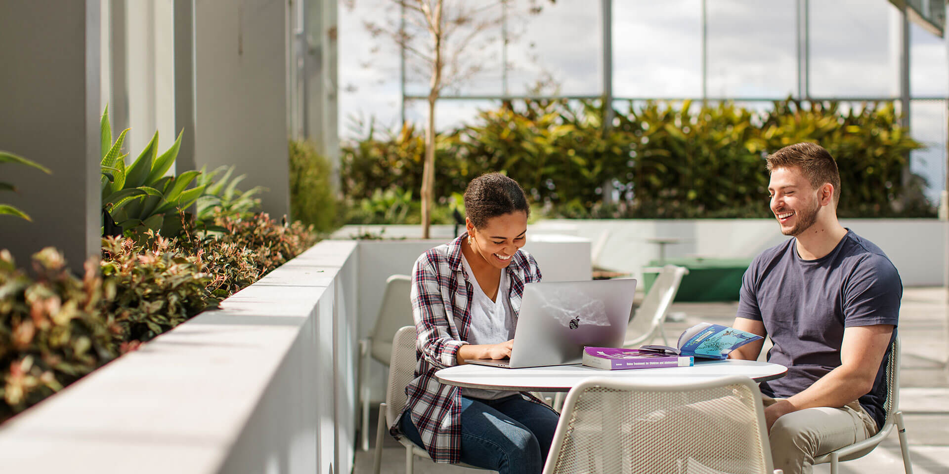 Two students sitting outdoors at a table studying with a laptop and textbooks
