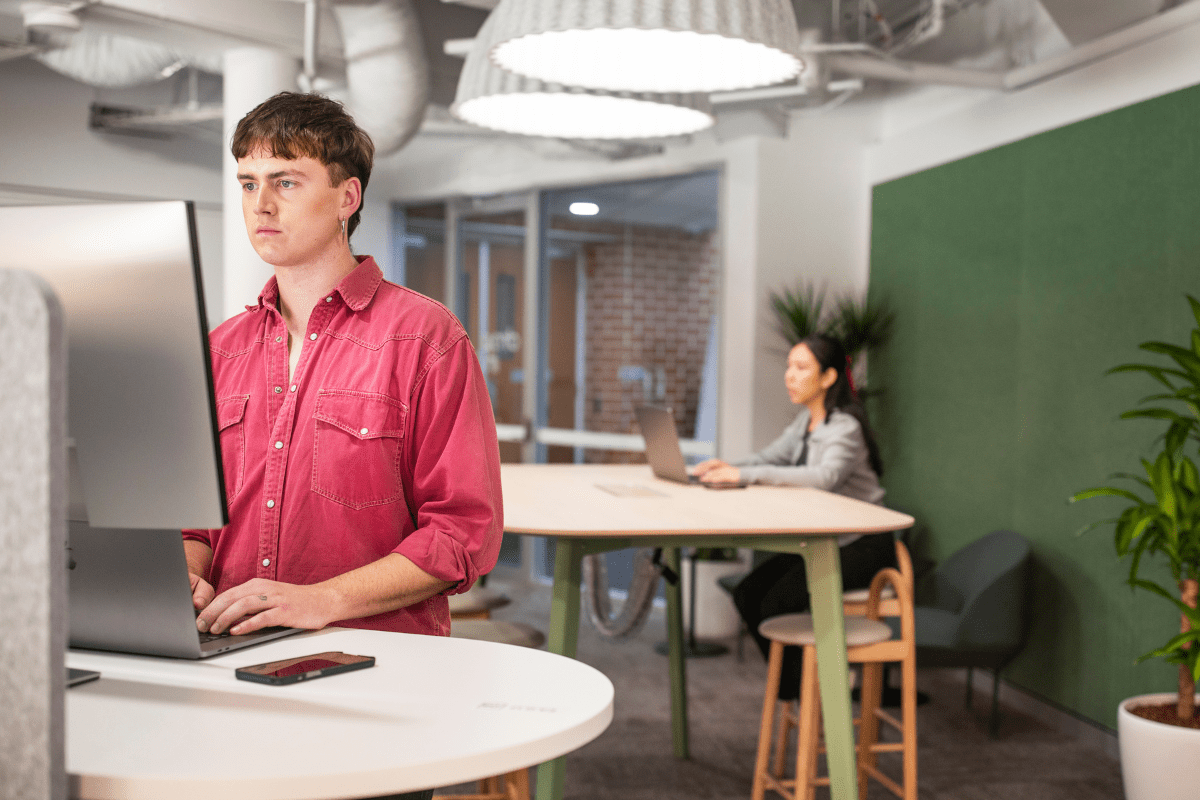 A male student is standing at a desk working on a computer. A female student can be seen in the background working on her laptop.