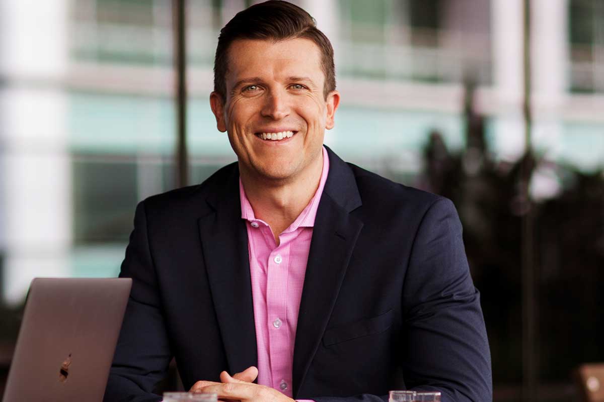 Male sitting outside wearing a pink collared shirt and dark blue suit jacket sitting and smiling at the camera