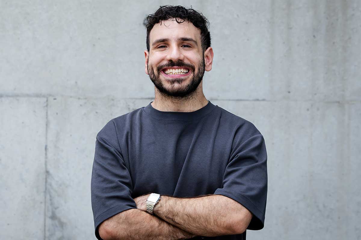 Male standing outside against a concrete wall wearing a dark grey shirt with arms crossed smiling at the camera