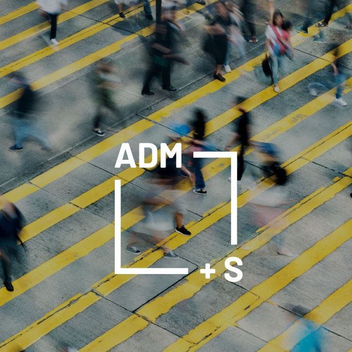 Approximately two dozen pedestrians walk in all directions across concrete which has yellow stripes painted diagonally across it. The figures are blurred and indistinct, indicating movement. The logo for the Centre for Excellence for Automated-Decision Making and Society is super-imposed on this image. The logo is a white box with the characters ADM+S on its perimeter.