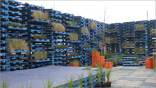 A pavilion made with blue wooden pallets. There are some plants in the pallets. Seating is in the form of orange plastic crates.