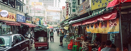 A street scene in Seoul, South Korea. The photo, taken from the middle of the street, shows a small truck diving away down the street, many shops and restaurants with people eating outdoors, and others walking on the street.